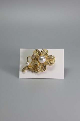 A gold-tone floral brooch with a central white pearl and a small pearl accent, displayed on a small white card over a textured white fabric background.