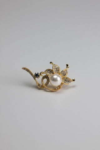 A gold-toned floral brooch with a central white pearl and small clear rhinestone accents, displayed on a small white card over a textured white fabric background.