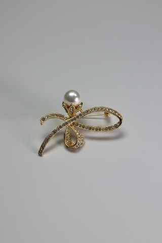 A gold bow-shaped brooch with a white pearl at the center and small clear stones along the edges, displayed on a small white card over a textured white fabric background.