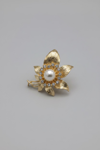 A gold-toned flower-shaped brooch with a central white pearl and small dark stones, displayed on a small white card against a textured white fabric background.