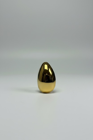 A polished gold teardrop-shaped brooch with a highly reflective, mirror-like surface, displayed on a small white card against a textured white background.