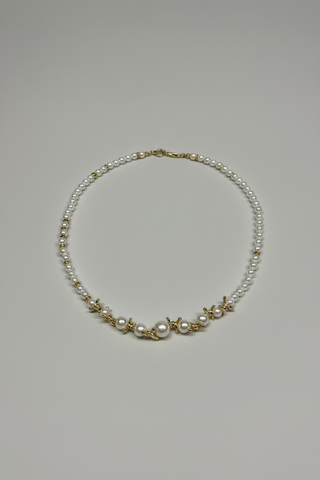 A delicate gold and pearl necklace laid flat on a textured white towel. The necklace features a strand of white pearls, small gold beads, and gold starfish charms.