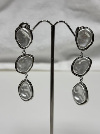 A pair of dangling earrings with irregular, baroque-shaped silver-toned beads, displayed on a black metal stand against a light gray fabric background.