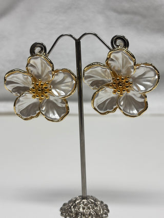 A pair of flower-shaped earrings with iridescent white petals and gold-tone centers, displayed on a silver-tone stand against a light gray background.
