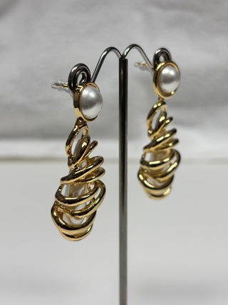 A pair of gold drop earrings with white pearl-like round studs, displayed on a black metal stand against a soft gray background.