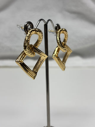 A pair of gold-toned geometric dangle earrings displayed on a black stand against a light gray fabric background.