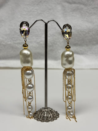 A pair of long dangling earrings displayed on a black jewelry stand against a light gray fabric background. Each earring has an iridescent oval stud, a gold bead, a large irregular white pearl, and multiple cascading pearl and gold chain drops.