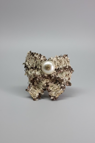 A vintage-style tweed brooch featuring a textured beige and brown bouclé pattern, a central white faux-pearl button, and a small gold star detail