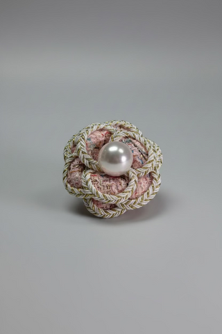 A decorative brooch featuring a central white pearl surrounded by a braided floral design in pink and gold