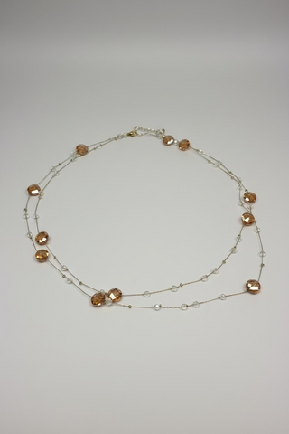 A delicate beaded necklace with faceted amber-orange beads and small clear beads on a thin silver-toned chain, laid out on a textured white fabric background.