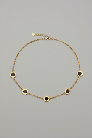 A gold chain necklace with five evenly spaced circular gold medallions, each featuring a dark black center, displayed on a neutral gray background.