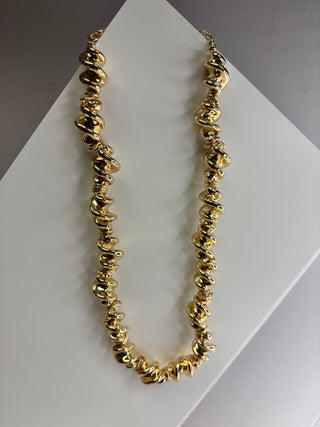 A chunky gold necklace with irregular, organic-shaped beads, displayed on a white geometric stand against a neutral gray background.