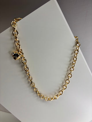 A gold chain necklace with a small heart-shaped charm featuring a black center, displayed on a white geometric surface.