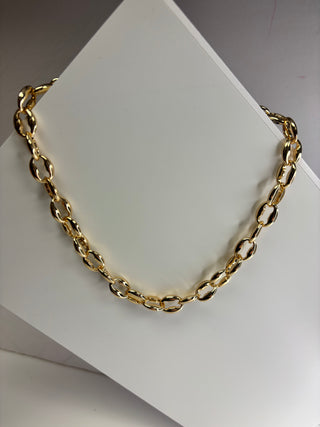 A gold-toned chain necklace with elongated oval links, draped on a white display stand against a neutral gray background.