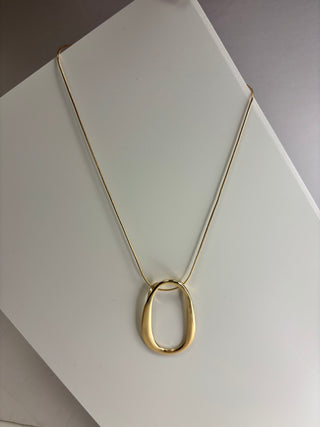 A gold pendant necklace with a thick, open oval loop hanging from a delicate gold chain, displayed on a white geometric surface.