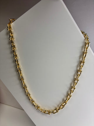 A gold-toned chain necklace with a textured, geometric link design, displayed on a white jewelry stand.