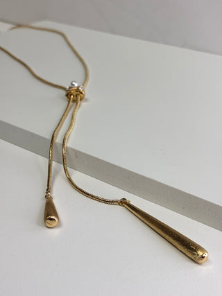 A gold-toned lariat-style necklace with a long snake-chain drape, a small knot detail, and two dangling teardrop-shaped pendants, displayed on a clean white surface.