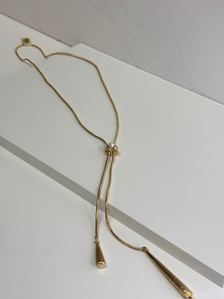 A gold-toned lariat-style necklace with a delicate chain, a central knot detail, and two dangling cone-shaped pendants, displayed on a clean white surface.
