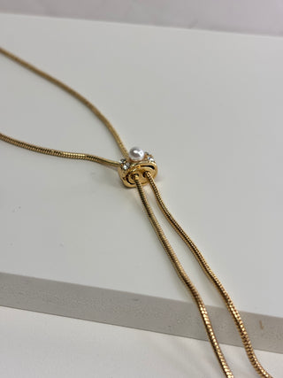 A gold snake chain necklace with a central knot-like clasp featuring a small white pearl and tiny clear stones, displayed on a light gray surface.