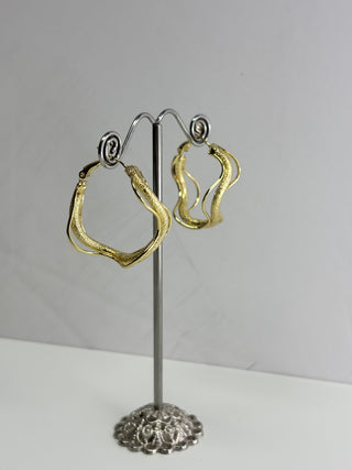 A pair of textured gold-tone hoop earrings displayed on a silver metal stand against a plain light gray background.
