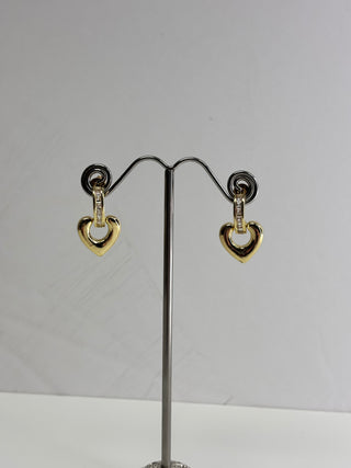 A pair of gold heart-shaped dangle earrings with small clear stone accents, displayed on a wire jewelry stand against a plain light gray background.